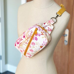 Puede incluir: Una ri&ntilde;onera blanca con estampado floral en tonos rosa y amarillo. La bolsa tiene una cremallera dorada y una correa ajustable de color amarillo mostaza.