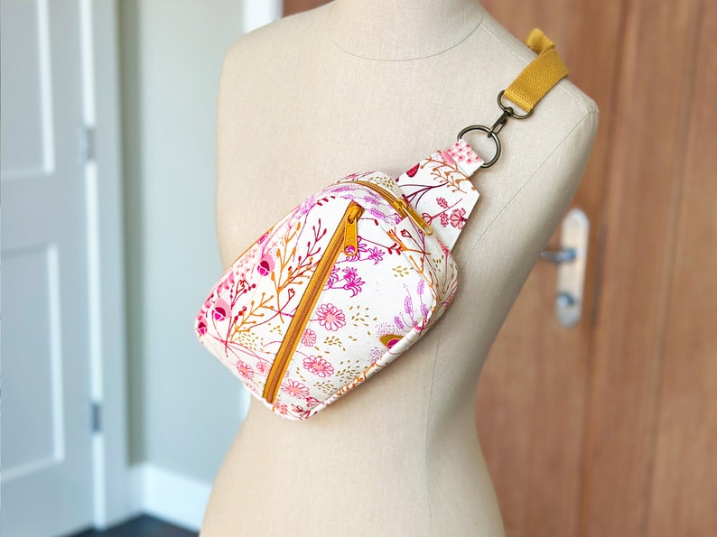Puede incluir: Una ri&ntilde;onera blanca con estampado floral en tonos rosa y amarillo. La bolsa tiene una cremallera dorada y una correa ajustable de color amarillo mostaza.