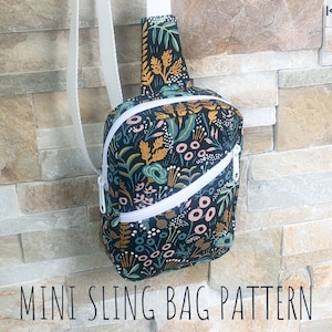 May include: A mini sling bag featuring a dark fabric with a colorful botanical print of leaves and flowers, a white diagonal zipper, and a white adjustable strap. The bag displays the text Kindred Sewls and Mini Sling Bag Pattern.