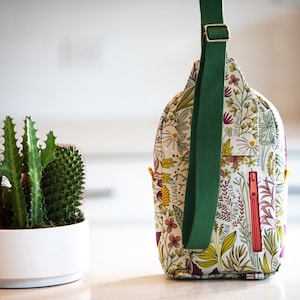 May include: A white and green floral print crossbody bag with a green adjustable strap. The bag has a zipper closure and a small pocket on the front.