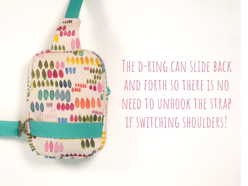 May include: A small crossbody bag with a cream-colored fabric featuring a multi-colored pattern of small painted shapes. It has a teal strap and a metal D-ring. Text reads: The D-ring can slide back and forth so there is no need to unhook the strap if switching shoulders!