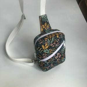May include: A crossbody bag featuring a black fabric base with a colorful botanical print of leaves, flowers, and stems in shades of yellow, teal, and pink. The bag includes a white zipper closure and an adjustable white woven strap.