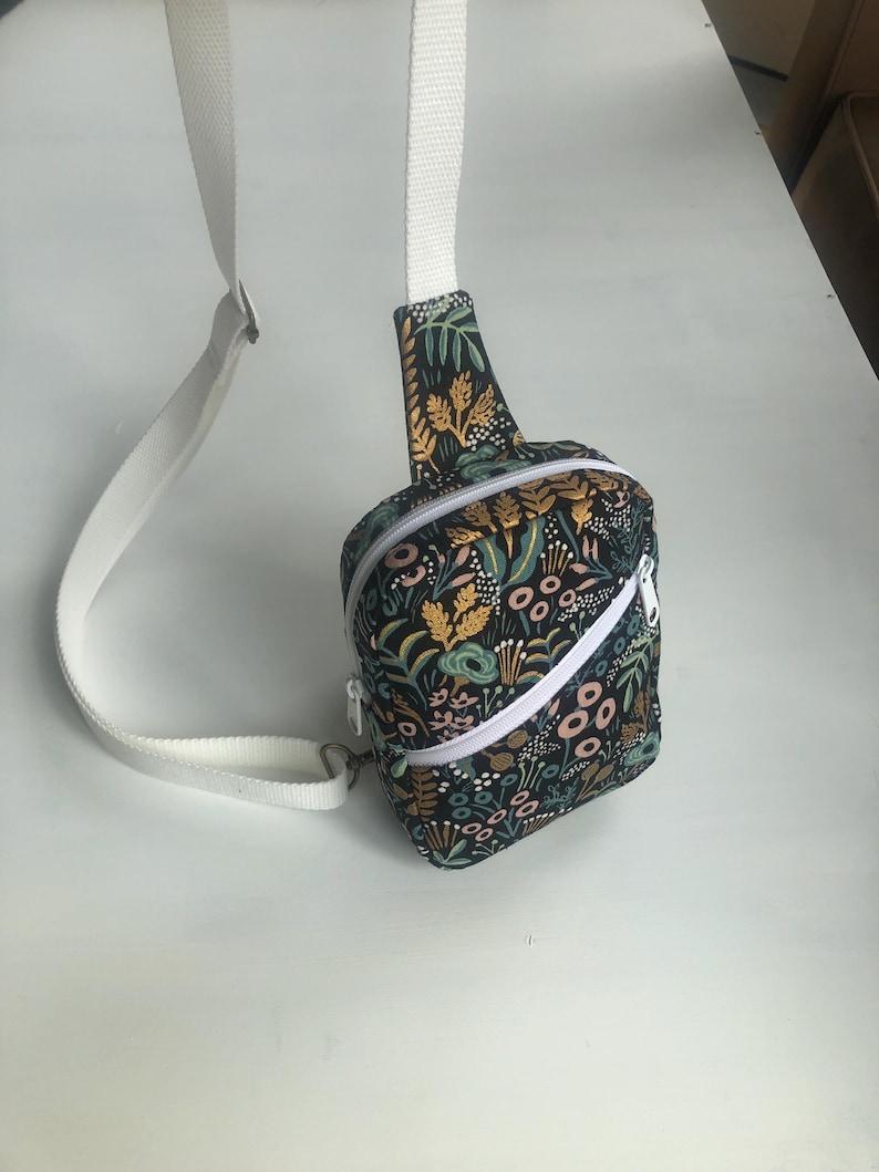 May include: A crossbody bag featuring a black fabric base with a colorful botanical print of leaves, flowers, and stems in shades of yellow, teal, and pink. The bag includes a white zipper closure and an adjustable white woven strap.