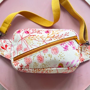 Puede incluir: Una peque&ntilde;a ri&ntilde;onera blanca con estampado floral en tonos rosa, rojo y dorado. La bolsa tiene una correa ajustable de color amarillo mostaza y una cremallera.