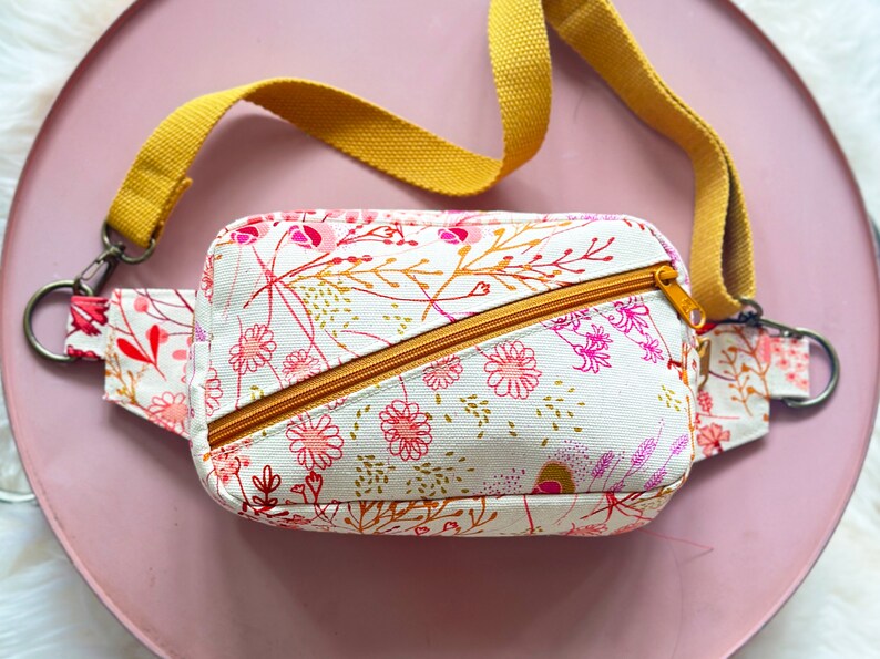 Puede incluir: Una peque&ntilde;a ri&ntilde;onera blanca con estampado floral en tonos rosa, rojo y dorado. La bolsa tiene una correa ajustable de color amarillo mostaza y una cremallera.