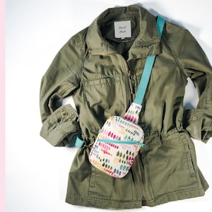 May include: A sling bag with a cream base and a colorful pattern of small, vertical, rounded shapes in shades of pink, yellow, green, and blue. The bag features a teal zipper and a matching teal strap. The bag is displayed over an olive green jacket.