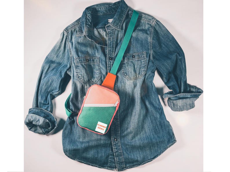 May include: A blue denim long-sleeve button-down shirt layered with a color-blocked crossbody sling bag. The bag features a peach top, a green bottom section, an orange strap, and a white rectangular label with the red text BE KINDER GENTLY.