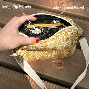 Puede incluir: Un bolso bandolera amarillo mostaza con un estampado floral blanco. El bolso tiene un bolsillo con cremallera y dos bolsillos deslizantes en el interior. El bolso tiene una correa de lona blanca.