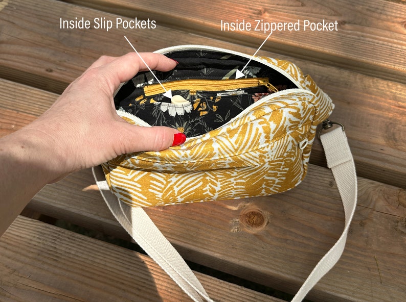 Puede incluir: Un bolso bandolera amarillo mostaza con un estampado floral blanco. El bolso tiene un bolsillo con cremallera y dos bolsillos deslizantes en el interior. El bolso tiene una correa de lona blanca.