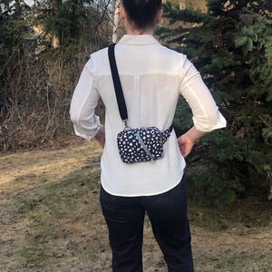 May include: A small rectangular crossbody bag featuring a black base with a colorful confetti-like pattern of white, pink, and teal shapes. The bag has a black adjustable strap, a silver zipper closure, and a diagonal silver reflective strip.