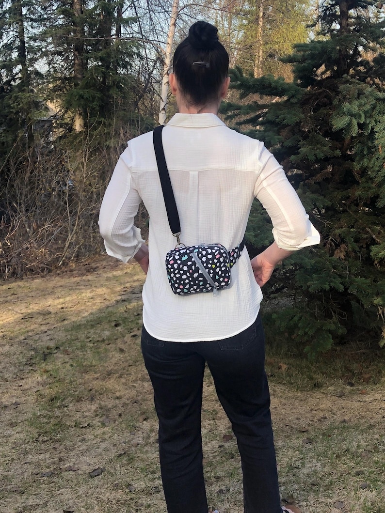 May include: A small rectangular crossbody bag featuring a black base with a colorful confetti-like pattern of white, pink, and teal shapes. The bag has a black adjustable strap, a silver zipper closure, and a diagonal silver reflective strip.