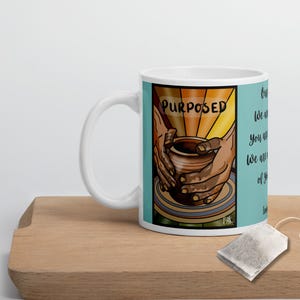 Può includere: Tazza in ceramica bianca con un'illustrazione colorata di una mano che modella l'argilla su un tornio da vasaio. Il testo "PURPOSED" è presente sulla tazza. La tazza è su una superficie di legno con una bustina di tè e un'etichetta gialla.