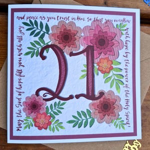 May include: A square birthday card featuring the number "21" in burgundy, framed by pink and orange floral designs and green foliage. The card includes text around the edges and is set against a textured white backdrop with a burgundy border. An envelope is visible.