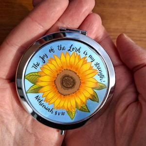 May include: A circular, silver compact mirror featuring a sunflower design. The sunflower has yellow petals and a brown centre, set against a light blue background. The text "The joy of the Lord is my strength! Nehemiah 8 v 10" is written around the edge.