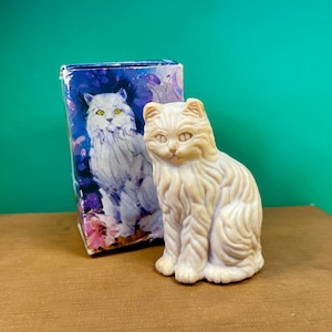 May include: A cream-colored, molded soap figurine shaped like a long-haired cat with detailed fur texture, positioned next to its rectangular cardboard packaging which features a colorful illustration of a white cat against a purple and blue background.