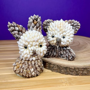 May include: Two seashell animal sculptures. One is a bunny with upright ears, the other a bear. Both are made of small, cream-coloured shells with brown accents. The bunny and bear have black eyes and are displayed on a wooden surface.