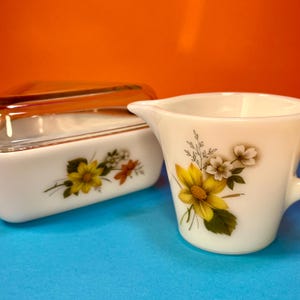 Könnte beinhalten: Ein weißes Keramik-Sahne- und Schüssel-Set mit Deckel. Beide Teile weisen ein Blumendesign mit gelben und weißen Blumen, grünen Blättern und braunen Akzenten auf. Der Sahnebereiter hat einen Griff und einen Ausguss. Die Schüssel hat einen Deckel aus klarem Glas.