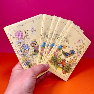 May include: A hand holds a stack of vintage greeting cards. Each card features a whimsical illustration of a mouse in a hat, surrounded by colourful flowers and foliage. The cards have a yellow background and a floral border.