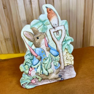 Peut inclure: Une figurine décorative représentant un lapin brun portant une veste bleue, tenant un radis rouge et se tenant à côté d'une pelle de jardin avec un petit oiseau brun et blanc perché au sommet, entouré de feuillage vert dans un jardin.