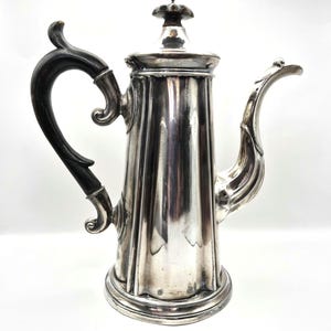 Puede incluir: Una cafetera plateada con un cuerpo cilíndrico alto y estriado, un pico curvo con detalles decorativos y un mango de madera oscura pulida. La tapa está rematada con un pequeño remate en forma de flor y la base es circular.