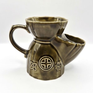 Op de afbeelding: Een olijfgroene keramische scheerkwast met een handvat en een ingebouwde zeepbak. De mok heeft reliëf bloemen- en kruisontwerpen. Het is ontworpen om scheerzeep en water te bevatten voor een traditionele natte scheerbeurt.