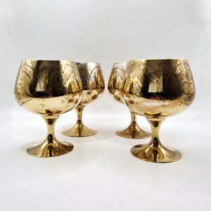 Könnte beinhalten: Vier goldfarbene Cognacschwenker mit geätzten floralen Designs. Die Gläser haben eine abgerundete Schalenform, einen schlanken Stiel und einen breiten Fuß. Die Gläser sind in einer Gruppe auf einer weißen Oberfläche angeordnet.