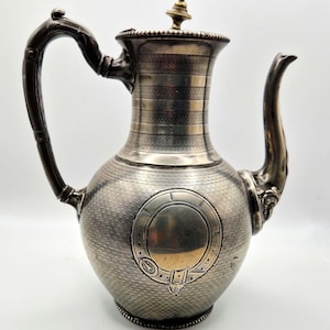 Op de afbeelding: Antieke verzilverde koffiepot met een getextureerde body en een decoratieve handgreep. De pot heeft een tuit, een deksel met een knop en een reliëf ontwerp aan de voorkant. Een vintage stuk voor het serveren van koffie of thee.