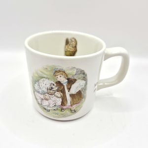 May include: A white ceramic mug featuring a colourful illustration of a hedgehog wearing a bonnet and a person in a brown coat. The mug has a handle on the right side and a small painted bird visible on the inner wall. The text is partially visible.