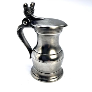 Könnte beinhalten: Ein kleiner Bierkrug in Zinnfarbe mit Klappdeckel und Henkel. Der Deckel ist mit einem dekorativen Element versehen. Der Krug hat einen bauchigen Körper mit horizontalen Bändern und einem gebogenen Henkel.