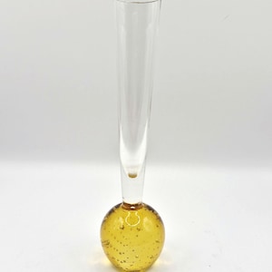 Op de afbeelding: Een heldere glazen bloemknopvaas met een lange, slanke hals en een ronde, amberkleurige basis. De basis is gevuld met kleine belletjes. De vaas is ontworpen om een enkele bloem of een klein bloemstuk te bevatten.