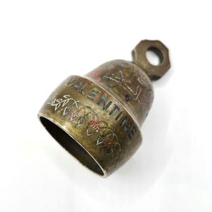 May include: A brass-toned metal bell featuring an etched design of a vine with heart-shaped leaves and the word VALENTINE engraved in capital letters. The bell has a dark, weathered patina and includes a small hexagonal loop at the top for hanging.
