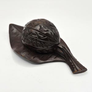 Peut inclure: Une boîte en bois sculpté brun foncé en forme de noix, posée sur une base en forme de feuille. La boîte en forme de noix présente des détails sculptés complexes, et la base en forme de feuille a un motif de veines détaillé. Le design est naturaliste et décoratif.