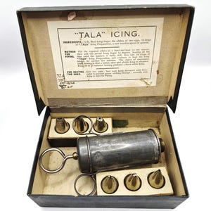 Può includere: Un antico set per glassare Tala in una scatola nera. Il set include una siringa per glassare in metallo con manico e diversi beccucci in ottone. La scatola contiene anche una ricetta stampata per la glassa Tala.