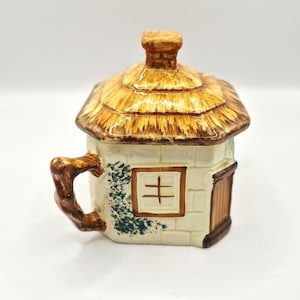 May include: A ceramic lidded jar shaped like a small cottage with a thatched roof, a chimney, a brown handle resembling a tree branch, a square window with brown trim, and a vertical door, featuring cream-coloured walls with green ivy detailing.