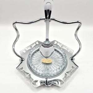 Op de afbeelding: Een zilverkleurige lepelhouder met een heldere glazen schaal en een lepel. De lepel rust in een houder boven de schaal. De schaal heeft een patroon van een sterrenuitbarsting. De lepelhouder is gemaakt van metaal en heeft een decoratief ontwerp.