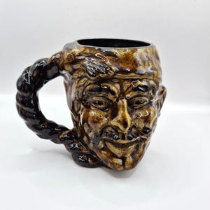 May include: A brown glazed ceramic mug shaped like a human face with a textured, rope-like handle. The mug features a glossy finish with dark brown highlights accentuating the facial features, including eyes, a nose, and a smiling mouth.