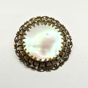 May include: A circular brooch featuring a central iridescent white mother-of-pearl disc surrounded by an ornate, antiqued bronze-toned metal frame with intricate filigree detailing and small, evenly spaced decorative metal tabs along the edge.