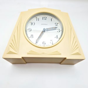 May include: Cream-coloured vintage Ingersoll clock with a geometric design. The clock face has black numerals and hands, with the brand name "Ingersoll" printed above the 6. The clock's design features a fan-like pattern on the sides.