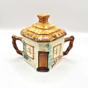 Può includere: Zuccheriera in ceramica a forma di casetta con tetto di paglia. La casetta ha un esterno color crema con finiture, finestre e una porta marroni. Il tetto e le maniglie sono marroni. Sono visibili accenti floreali verdi e rossi.