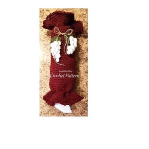 May include: A burgundy crochet wine bottle bag with a ruffled top and bottom. The bag is decorated with white and green grape clusters and a twine bow. The text "NeedleMeThat Crochet Pattern" is visible.