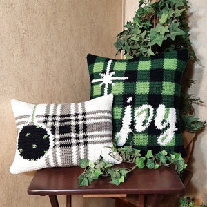 May include: Two crocheted throw pillows with a green and black plaid pattern. One pillow has the word "joy" written in white on it. The other pillow has a black and white plaid pattern with a green and black ornament.