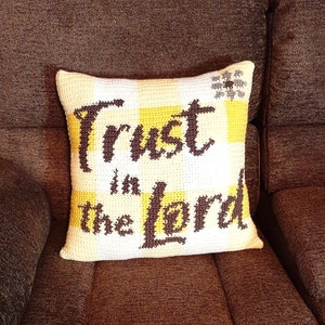 May include: A white crocheted pillow with a yellow and white checkered pattern. The pillow has the words "Trust in the Lord" embroidered in brown on it.