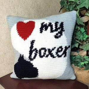 May include: A crocheted pillow with a white background and black and red accents. The pillow features a red heart, a black paw print, and the text "I love my boxer".