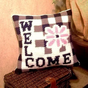 May include: A black and white checkered pillow with the word "WELCOME" in white letters and a pink flower in the center. The pillow is sitting on a brown wicker basket.