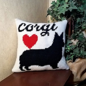 May include: White crocheted pillow with a black silhouette of a corgi dog and a red heart. The pillow has the word "corgi" embroidered on it in black.