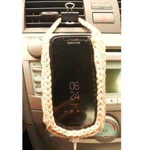May include: A beige crocheted phone holder with a clip attached to the top. A black smartphone is held in the holder and plugged into a charging cable. The phone is in a car with the car's dashboard and air vents visible in the background.