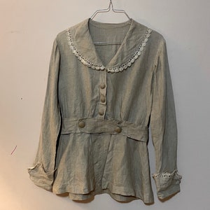 May include: A light gray linen blouse with a Peter Pan collar trimmed with white lace. The long-sleeved top features a button-down front, a cinched waist with a belt detail, and lace cuffs. The blouse is hanging on a metal hanger.