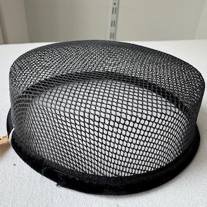 May include: A black mesh hat with a rounded crown and a black velvet band around the base. The hat has a tag attached to it. The hat is likely vintage or a costume piece.