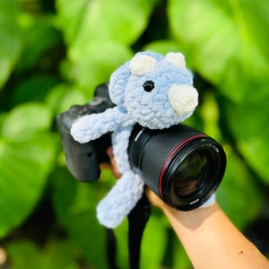 May include: A blue crocheted triceratops dinosaur toy is attached to a black camera lens with a red ring. The camera is held by a hand.