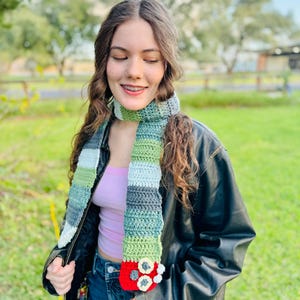 May include: A handmade crochet scarf in shades of green, gray, and white, with a red and white flower design at the end. The scarf is worn over a black leather jacket and blue jeans.